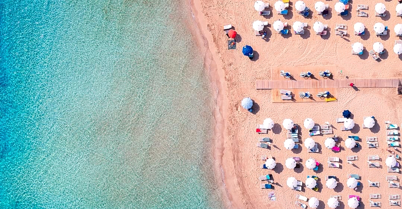 Fig Tree Bay in Protaras from above: dense rows of sunbeds and umbrellas on fine golden sand beside clear turquoise shallows on the Blue Flag beach