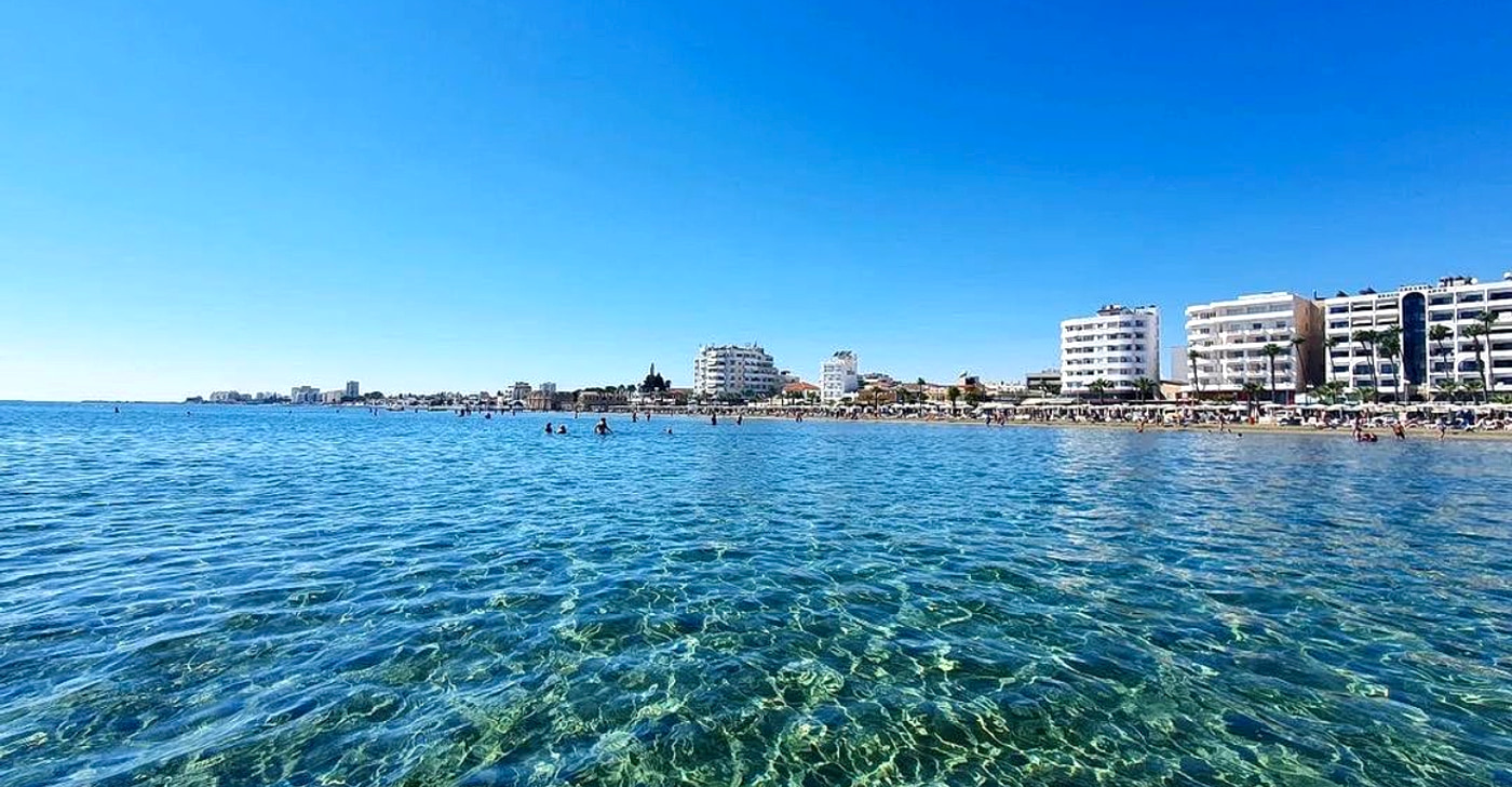 Finikoudes beach in Larnaca seen from the water: clear shallow sea in the foreground and the city seafront apartment blocks lining the beach behind