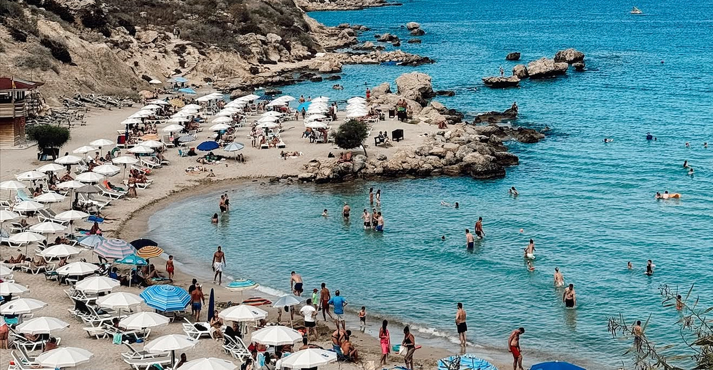 Konnos Bay between Ayia Napa and Protaras: a sheltered cove of golden sand backed by scrub-covered slopes, with umbrella rows above calm turquoise water and a boat anchored offshore