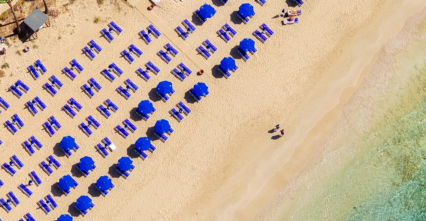Mackenzie Beach in Larnaca from above: blue umbrellas and sunbeds lined up in tight rows on golden sand beside clear turquoise shallows